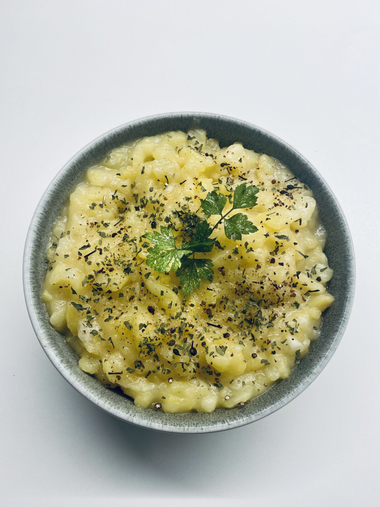 Eine Schüssel cremiges Risotto, bestreut mit Kräutern und garniert mit einem Zweig frischer Petersilie, erinnert vor einem weißen Hintergrund an die reichen Aromen der traditionellen schwäbischen Küche.