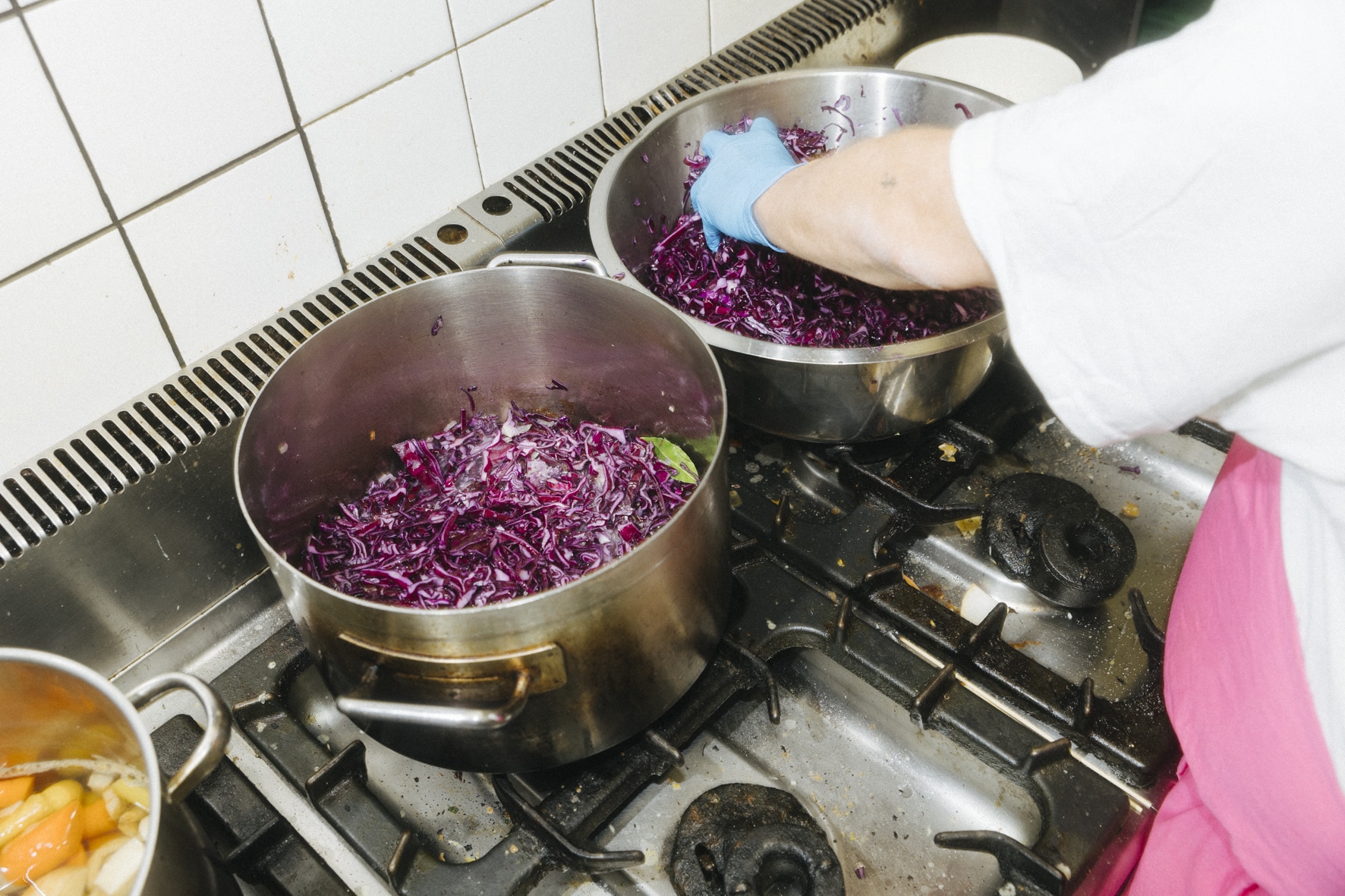 Eine Person in weißem Hemd und rosa Schürze rührt zerkleinerten Rotkohl in einem großen Topf auf dem Herd um und vermischt die Aromen mit Gojuang. Daneben steht ein weiterer Topf voller Gemüse.