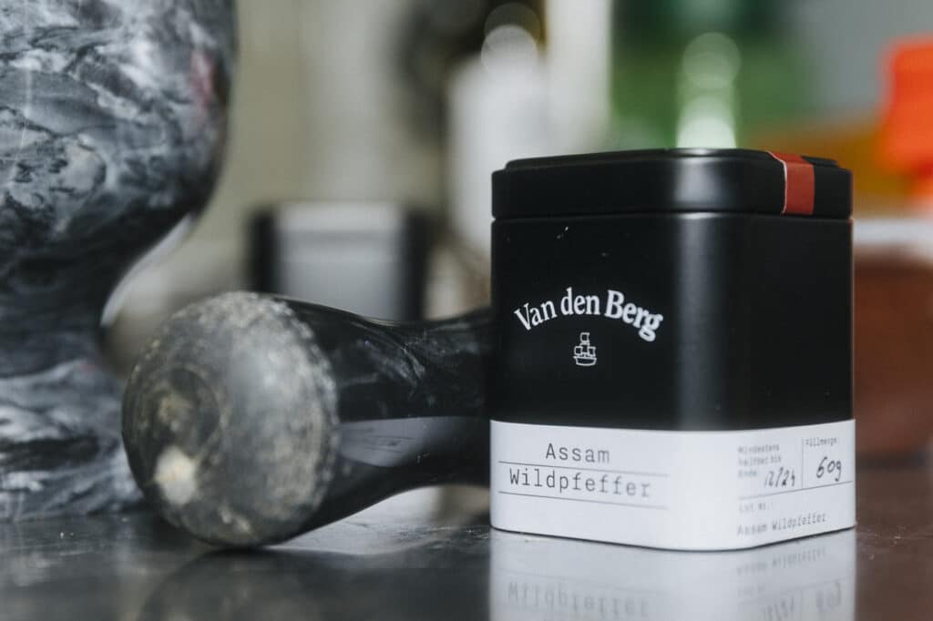 Eine Metalldose mit der Aufschrift „Van den Berg Assam Wildpepper“ steht auf einer reflektierenden Oberfläche neben einem dunklen, runden Objekt, das an einen Kung-Fu-Trainingsball erinnert. Die Kulisse scheint eine Küche oder ein Arbeitsplatz mit verschwommenen Gegenständen im Hintergrund zu sein, was eine geheimnisvolle Atmosphäre wie bei einem Gansl-Festmahl verleiht.