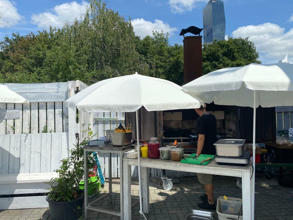 Eine Person steht unter weißen Schirmen an einer Außenküche und bereitet sich auf großartiges Kochen vor. Auf dem Tisch sind verschiedene Vorräte ausgebreitet. In der Nähe wartet ein Grill auf seinen Einsatz, während Bäume und ein hohes Gebäude in den teilweise bewölkten Himmel ragen und die perfekte Kulisse für jeden hungrigen kulinarischen Enthusiasten bieten.