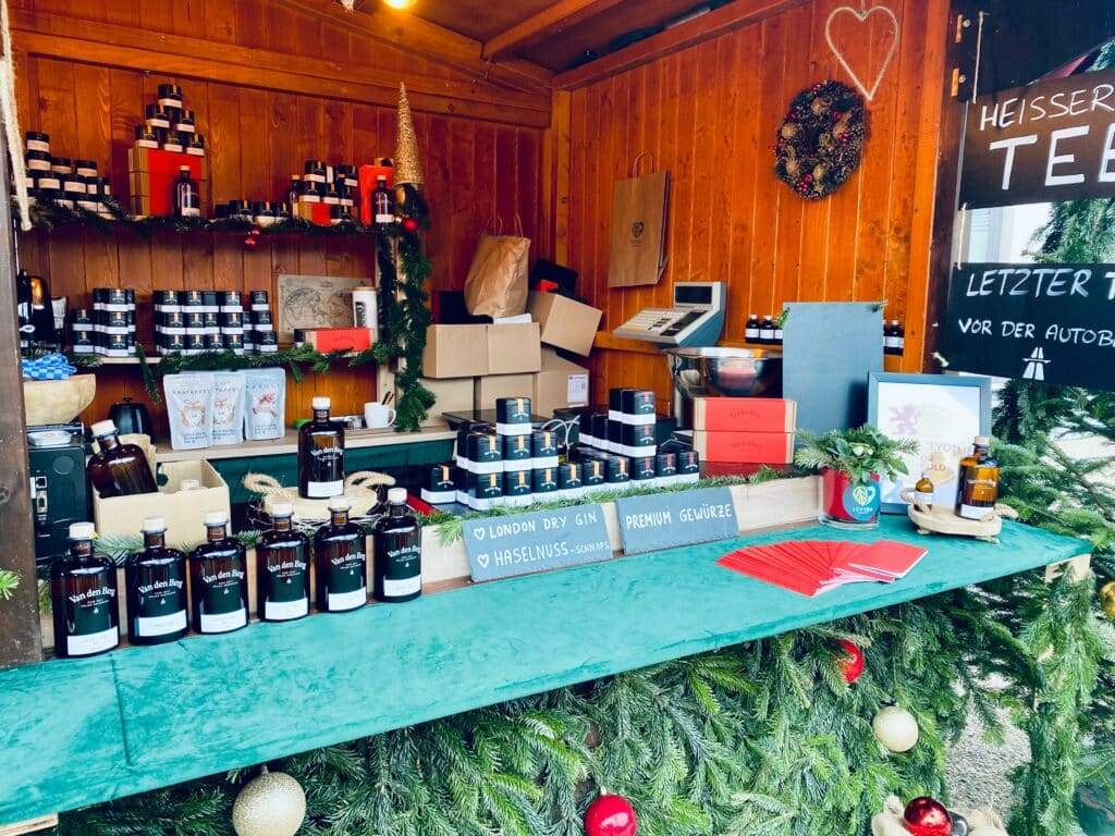 Ein festlicher Marktstand präsentiert Van den Bergs Gin mit Haselnussgeschmack, umgeben von Weihnachtsdekorationen. Im Hintergrund sind eine Registrierkasse und Stapel von Kartons zu sehen. Tannenzweige und Ornamente schmücken den Holzstand und verleihen ihm einen weihnachtlichen Charme.