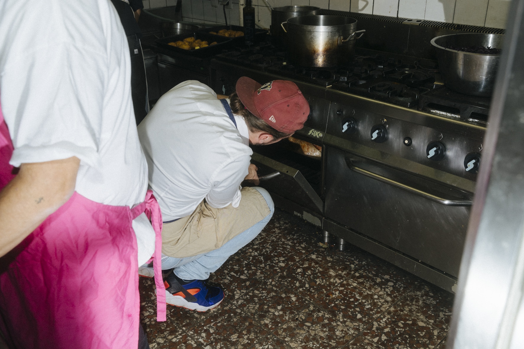 Eine Person mit roter Mütze und Schürze überprüft in einer geschäftigen Küche mit der Präzision des Kung Fu das Essen in einem Industrieofen. Eine andere Person in einer rosa Schürze, die möglicherweise Gansl zubereitet, ist teilweise zu sehen und steht in der Nähe. Die Küche ist voll mit großen Töpfen und verschiedenen Kochutensilien.