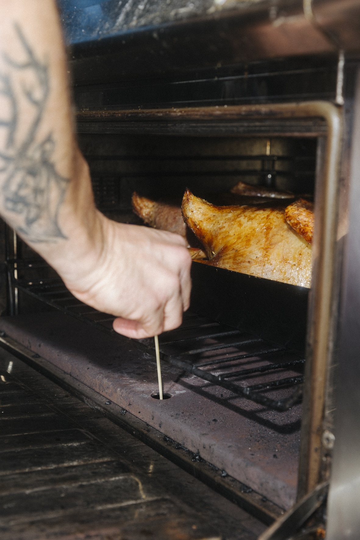 Eine Person mit tätowierter Hand prüft gekonnt mit einem Metallspieß die Temperatur eines gebratenen Gansl im Ofen. Das Huhn sieht goldbraun und knusprig aus.
