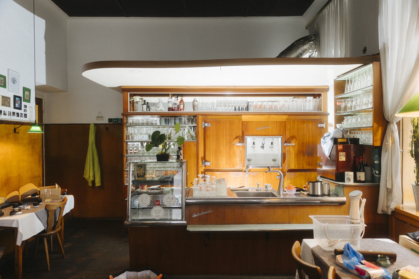 Ein gemütliches Café-Interieur im Vintage-Stil mit Holzvertäfelung, einer Theke mit Gläsern und Spüle sowie einer Speisekarte mit Gansl-Spezialitäten. Tische mit weißen Tischdecken und Stühlen stehen bereit und durch das Fenster fällt sanftes Tageslicht. Auf der Theke steht eine Pflanze.