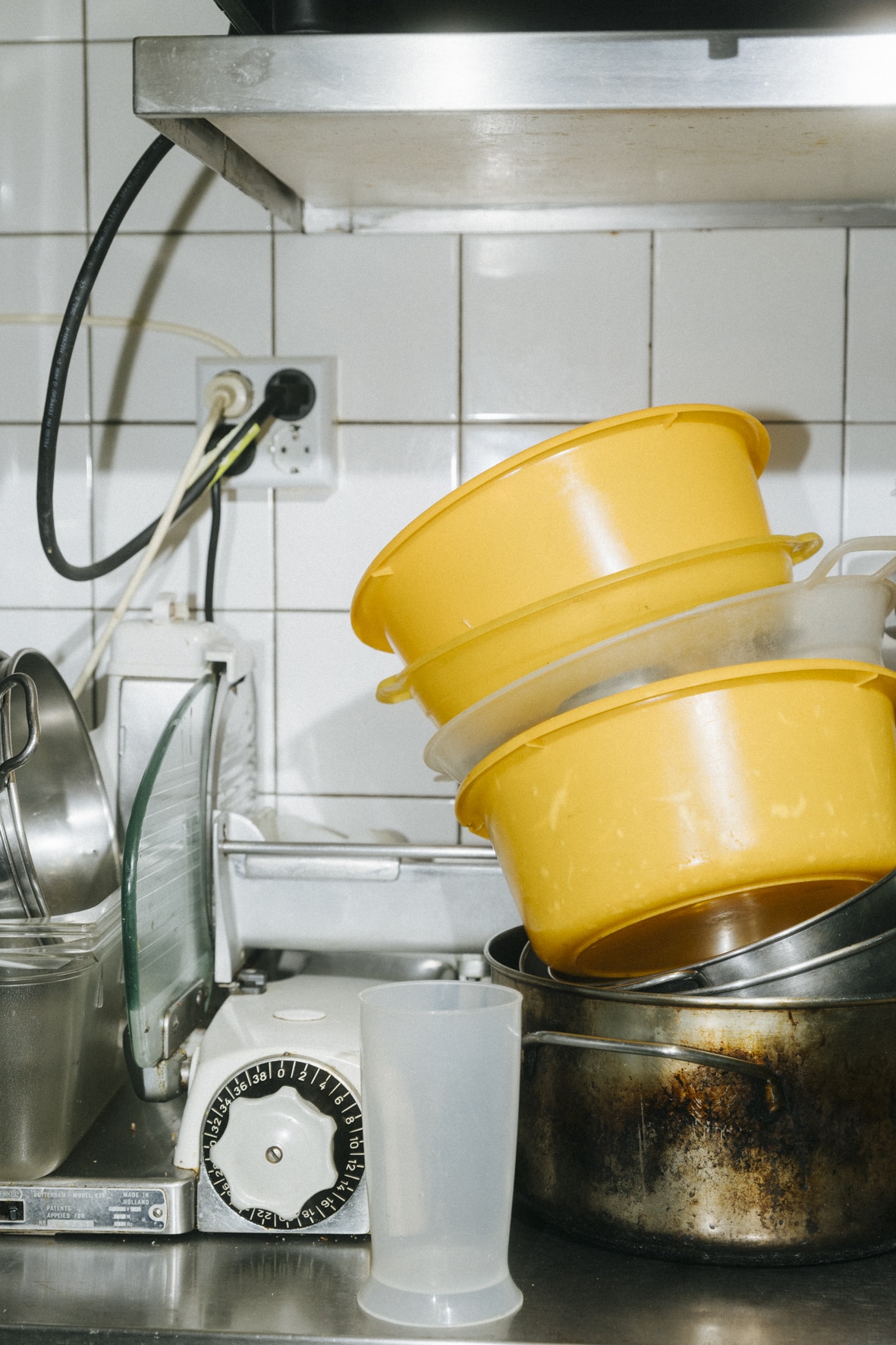 Eine Küche mit gestapelten gelben Plastikschüsseln, einem Metalltopf und einer durchsichtigen Tasse auf der Theke erinnert an einen Kung-Fu-Gansil-Trainingsplatz. In der Nähe vor den weiß gefliesten Wänden steht ein Küchenwecker. Über der Theke ist eine einsatzbereite Steckdose zu sehen.