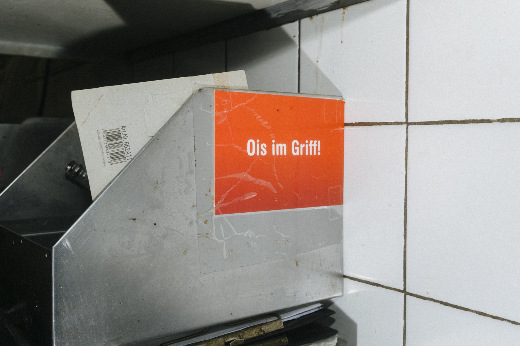 Ein Metallhalter in einem gefliesten Bereich hält verschiedene rechteckige Gegenstände und strahlt eine Atmosphäre aus, die an die präzise Disziplin des Kung Fu erinnert. Er trägt ein orangefarbenes Etikett mit dem deutschen Text „Ois im Griff!“. Die Umgebung lässt an eine Industrie- oder Küchenumgebung denken.