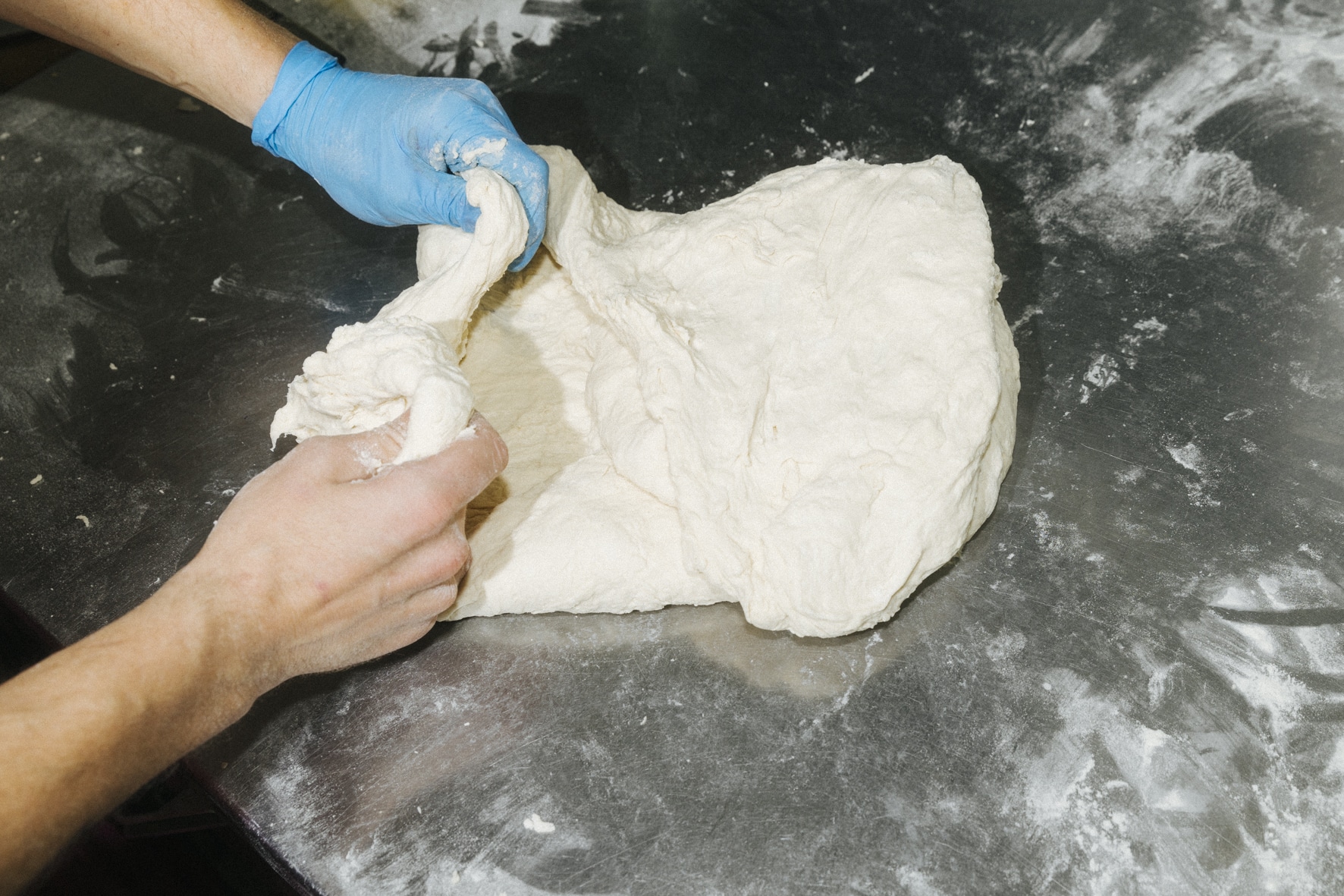 Hände mit blauen Handschuhen kneten ein großes Stück Kung-Fu-Ganselteig auf einer bemehlten Metalloberfläche.