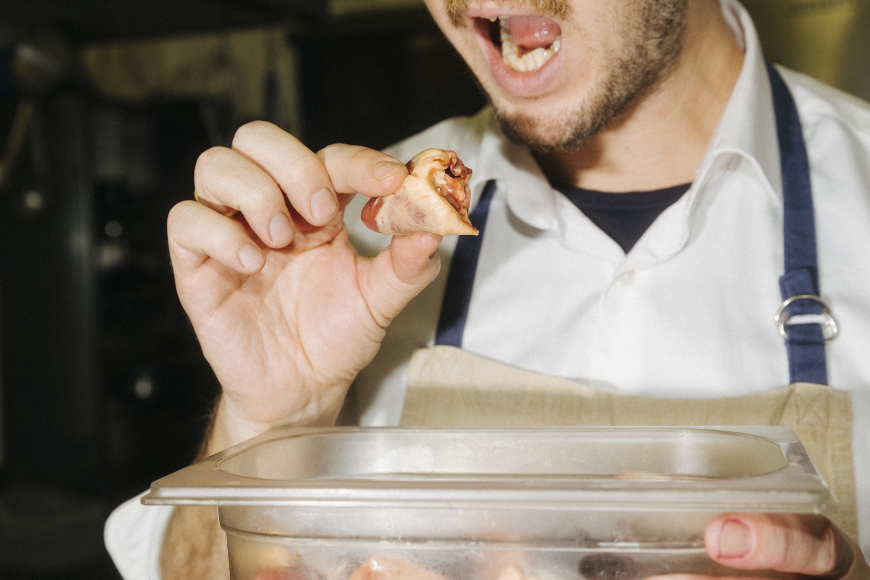 Eine Person in weißem Hemd und Schürze hält mit der Präzision des Kung Fu ein Stück Essen an den Mund. Im Vordergrund ist ein Behälter mit ähnlichen Essensstücken zu sehen, was darauf schließen lässt, dass die Person diese Kochkunst gemeistert hat. Die Szene scheint eine Küche zu sein.